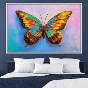May include: A framed canvas art piece featuring a vibrant butterfly painting. The butterfly has colorful wings with shades of yellow, orange, blue, and brown. The background has a blend of blue, purple, and pink hues.