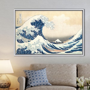 May include: Framed print of Hokusai's "The Great Wave off Kanagawa." The artwork depicts a large, stylized wave with white foam and blue water, boats, and a mountain in the background. The print is in a white frame and hangs on a light gray wall.