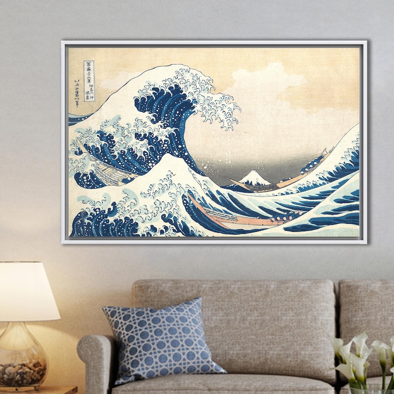 May include: Framed print of Hokusai's "The Great Wave off Kanagawa." The artwork depicts a large, stylized wave with white foam and blue water, boats, and a mountain in the background. The print is in a white frame and hangs on a light gray wall.