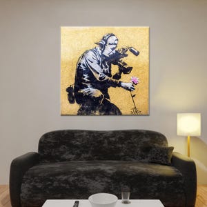 Puede incluir: Una impresión de arte de esténcil en blanco y negro de una persona sosteniendo una cámara y una flor. El fondo es de color amarillo dorado.