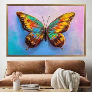 May include: A framed butterfly painting with a vibrant color palette. The butterfly features wings in shades of yellow, orange, blue, and brown, set against a background of blue and pink hues. The artwork is displayed in a gold-colored frame.