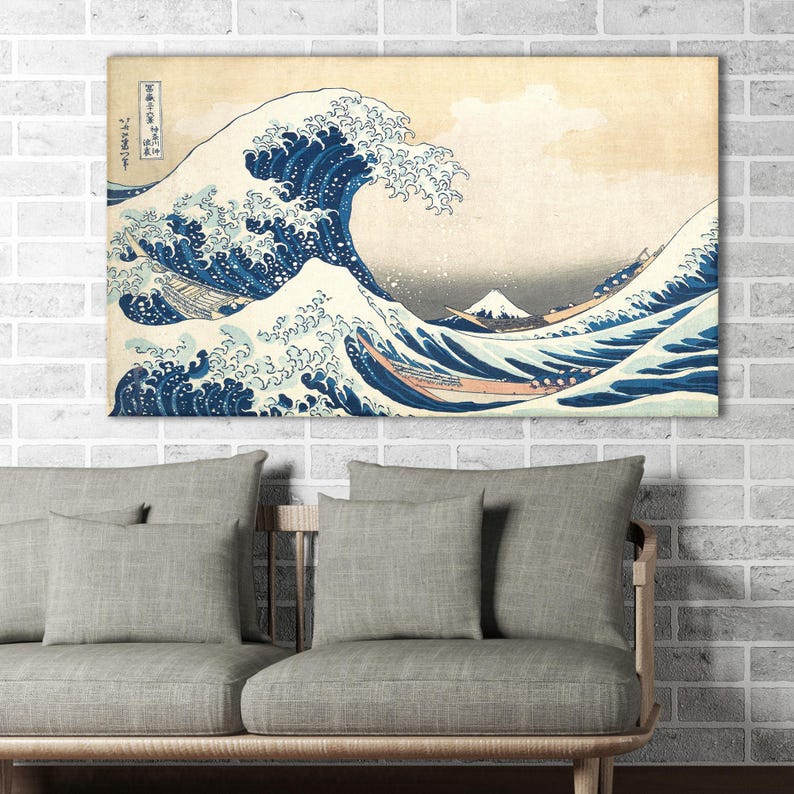 May include: A framed print of Hokusai's "The Great Wave off Kanagawa" features a large, curling wave in shades of blue and white, with boats and Mount Fuji in the background. The artwork is displayed above a gray sofa with matching pillows.