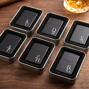 May include: Six matte black metal lighters, each engraved with a unique two-letter monogram, are presented in individual silver-toned metal tins with black foam inserts. The monograms include KG, DR, SN, AE, ZH, and BG in a serif font.