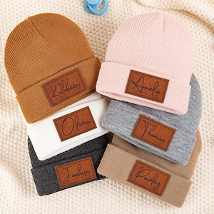 Pode incluir: Uma coleção de seis gorros de bebé personalizados em várias cores, incluindo castanho, rosa, cinzento, branco e bege. Cada gorro apresenta um remendo de couro castanho com um nome em escrita cursiva. Os gorros estão dispostos sobre um cobertor creme.