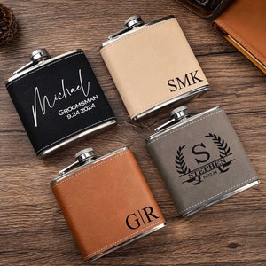 May include: Four personalized leather-wrapped hip flasks with different engravings. The flasks are in various colors, including black, brown, beige, and gray. The engravings include names, dates, and initials.
