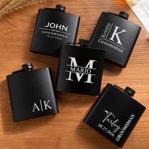 May include: Five matte black metal flasks with screw-top caps, each featuring unique white laser-engraved personalisation. The designs include names like John, Kristin, Mario, and Tarloy, along with dates and initials, arranged in a flat lay composition.