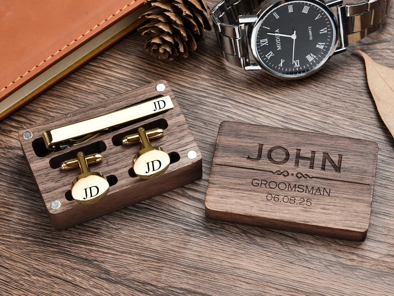 May include: A wooden box with a gold tie clip and cufflinks. The box is engraved with the name "JOHN" and the words "GROOMSMAN" and the date "06.08.25".