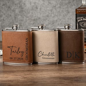 May include: Three metal flasks wrapped in textured faux leather in tan, beige, and dark brown. Each flask features a silver screw-top lid and custom laser-engraved text, including names like Tarloy and Charlotte, dates, and the word Groomsman.