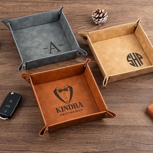 Può includere: Tre vassoietti in pelle personalizzati, grigio, beige e marrone. Il vassoio grigio ha la lettera "A", quello beige le iniziali "SHP", e quello marrone una grafica di smoking con la scritta "KINDRA GROOMSMAN".