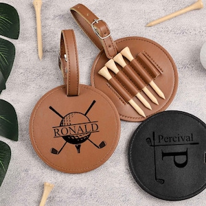 May include: Two personalized leather golf bag tags. One is brown with a golf ball and crossed clubs design and the name "RONALD" engraved. The other is black with a golf club and the initial "P" engraved and the name "Percival" above it. Both tags have a leather strap.