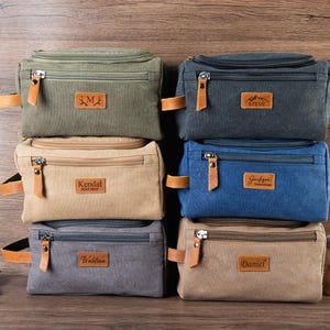 Op de afbeelding: Zes canvas toilettassen in verschillende kleuren, gestapeld. Olijfgroen, marineblauw, beige, blauw, grijs en beige. Elke tas heeft een ritssluiting, lederen details en een gepersonaliseerd leren label. Een scheerkwast en een klein blikje zijn zichtbaar.