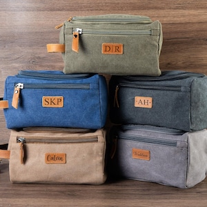 May include: A collection of canvas wash bags in shades of green, blue, grey and beige. Each bag features a zip, leather details, and a personalised patch with initials. A shaving brush and accessories are visible in the background.