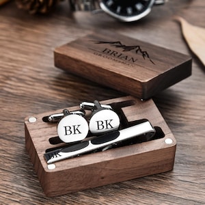 May include: A wooden box with a personalized engraved message "Brian Groomsman" and a mountain design. The box contains a tie clip and cufflinks with the initials "BK" engraved on them.