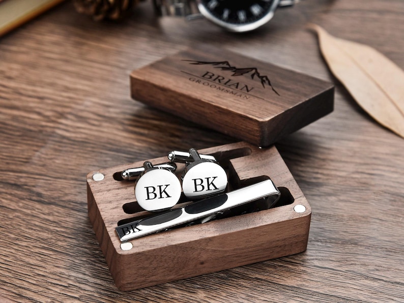 May include: A wooden box with a personalized engraved message "Brian Groomsman" and a mountain design. The box contains a tie clip and cufflinks with the initials "BK" engraved on them.