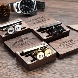 May include: Six wooden boxes with engraved names and dates, each containing a tie clip and cufflinks. The boxes are arranged in a row on a wooden surface.