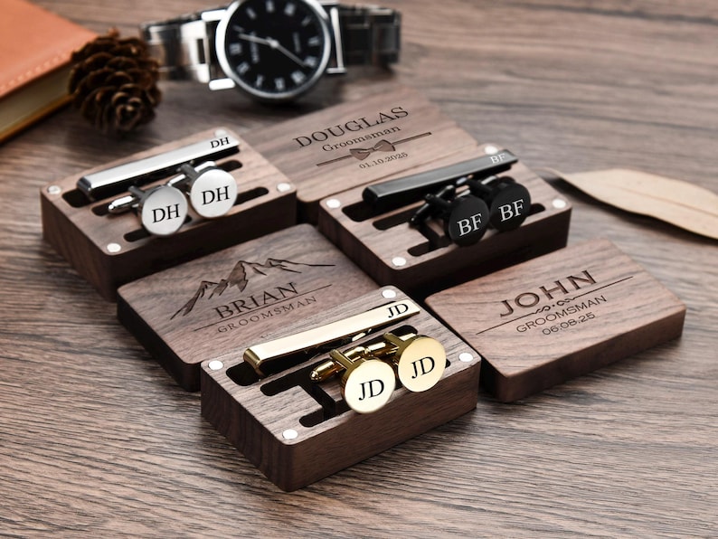 May include: Six wooden boxes with engraved names and dates, each containing a tie clip and cufflinks. The boxes are arranged in a row on a wooden surface.