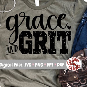 Peut inclure: Un t-shirt vert avec le texte "grace and grit" en noir avec un aspect vieilli. Le texte est en écriture cursive. Le t-shirt est posé sur une table avec un jean et une botte en cuir marron.