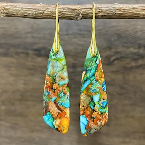 Peut inclure: Paire de boucles d'oreilles pendantes avec des crochets dorés et des pierres en forme de goutte. Les pierres présentent une mosaïque de turquoise, d'orange, de vert et de marron. Les boucles d'oreilles sont présentées sur un fond en bois flou.