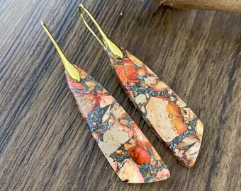 May include: A pair of dangle earrings with gold hooks and a long, teardrop-shaped stone. The stone is a mix of beige, orange, and black colors, resembling a marbled pattern.