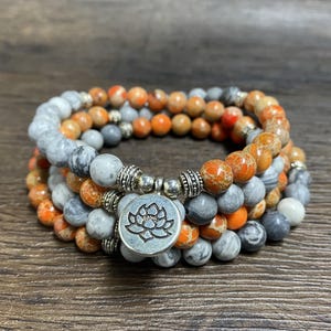 Könnte beinhalten: Ein dreifach gewickeltes Armband mit grauen und orangefarbenen Steinen und einem silbernen Anhänger mit einem Lotusblumenmotiv.