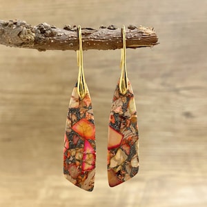 May include: Dangle earrings with gold-coloured hooks and teardrop-shaped stone pendants. The stone has a mosaic of pink, orange, and beige, with dark brown accents. The earrings are displayed on a small wooden branch.