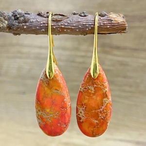 May include: A pair of dangle earrings featuring teardrop-shaped orange and white jasper stones. The earrings have gold-toned hooks.