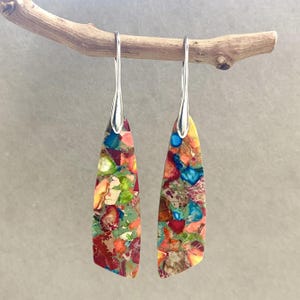 Puede incluir: Un par de pendientes colgantes con ganchos de plata y gotas de piedra multicolores y multifacéticas. Las piedras son una mezcla de rojo, naranja, amarillo, verde, azul y marrón.