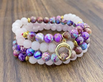 Puede incluir: Tres pulseras superpuestas con cuentas de piedras preciosas rosas, moradas y azules. La capa superior tiene un dije de media luna de oro.