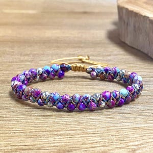 May include: A purple, blue, and gold beaded bracelet with a woven design. The bracelet has a gold clasp.