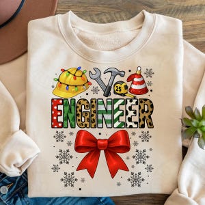 May include: A cream-colored sweatshirt with a festive design. The word "ENGINEER" is displayed in a mix of patterns and colors, with a red bow at the bottom. Above the word are engineer-related icons and snowflakes.