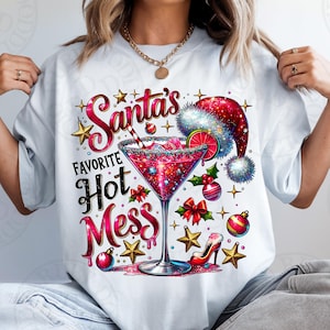Może przedstawiać: Biała koszulka z kolorowym wzorem graficznym przedstawiającym szklankę martini z czerwono-białym kapeluszem Świętego Mikołaja, czerwono-zielonym ostrokrzewem i tekstem "Santa's Favorite Hot Mess".