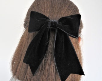 May include: A large black velvet hair bow featuring a structured center knot and two long, wide ribbon tails that drape downwards. The fabric has a soft, light-reflecting texture characteristic of velvet, secured to the hair with a clip.