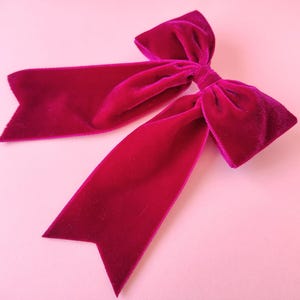 K&ouml;nnte beinhalten: Eine leuchtend magentafarbene Samtschleife mit langen, spitz zulaufenden Enden. Die Schleife besteht aus einem weichen, pl&uuml;schigen Material und ist vor einem hellrosa Hintergrund platziert. Diese dekorative Schleife eignet sich f&uuml;r Haarschmuck oder Geschenkverpackungen.