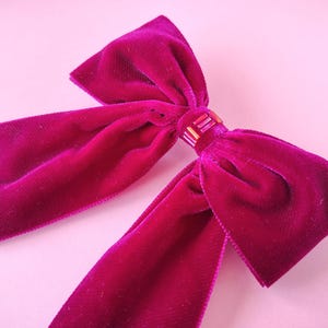 K&ouml;nnte beinhalten: Nahaufnahme einer magentafarbenen Samtschleife mit langen, flie&szlig;enden Enden. Die Schleife ist in der Mitte mit einer kleinen, rechteckigen Verzierung mit einer Mischung aus goldenen und rosa Perlen gebunden. Der Stoff hat eine weiche, pl&uuml;schige Textur.