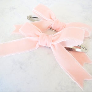 Puede incluir: Dos pinzas para el pelo de terciopelo rosa con clips de metal plateado. Los lazos est&aacute;n atados con un ribete blanco.