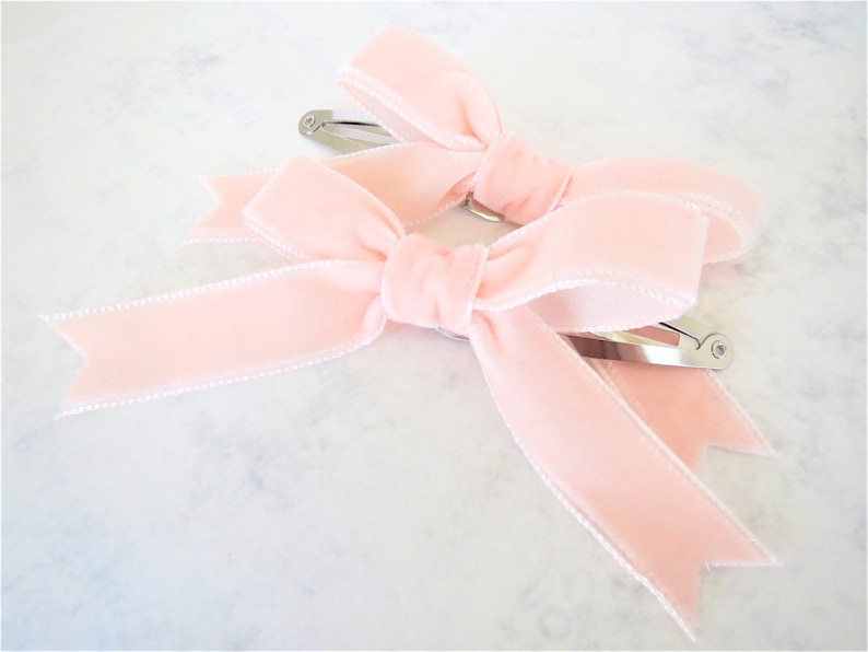 Puede incluir: Dos pinzas para el pelo de terciopelo rosa con clips de metal plateado. Los lazos est&aacute;n atados con un ribete blanco.