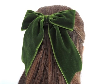 May include: A dark forest green velvet hair bow featuring a classic tied knot center and two long, wide fabric tails. The soft, plush texture of the velvet material catches the light, creating subtle highlights across the surface of the bow.