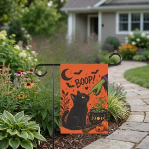 Könnte beinhalten: Eine rechteckige Gartenflagge mit orangefarbenem Hintergrund, die eine schwarze Katze, eine grüne Hand, die die Nase der Katze berührt, und den Text BOOP zeigt. Das Design umfasst eine Mondsichel, Fledermäuse, einen Kessel mit grünem Rauch und zwei Kürbisse.