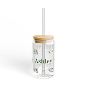 Peut inclure: Un gobelet en verre transparent avec un couvercle en bambou et une paille en verre. L'extérieur présente un motif répétitif de tuiles de mah-jong et le prénom Ashley imprimé en vert foncé. Le design inclut des accents rouges et verts.