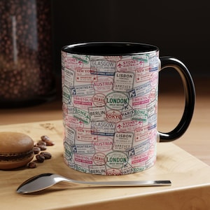 May include: A white ceramic mug with a black interior and handle, featuring a repeating pattern of colorful international travel passport stamps from cities including London, Paris, Berlin, Tokyo, New York, and Sydney, along with various dates and airport codes.
