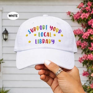 Peut inclure: Casquette de baseball blanche avec le texte "SUPPORT YOUR LOCAL LIBRARY" en couleurs arc-en-ciel. La casquette a une visière incurvée et est faite d'un matériau souple. Le texte est brodé avec des étoiles jaunes.