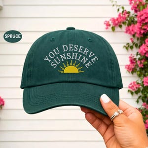 Può includere: Cappellino da baseball verde scuro con la scritta ricamata "YOU DESERVE SUNSHINE" in bianco e un sole giallo. Il cappellino ha un classico design a sei pannelli con visiera curva.