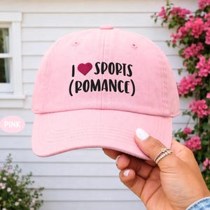 May include: A light pink baseball cap featuring the embroidered black text I HEART SPORTS (ROMANCE) with a small red heart icon replacing the word heart. The cap has a structured crown, a curved brim, and stitched eyelets on the top panels.