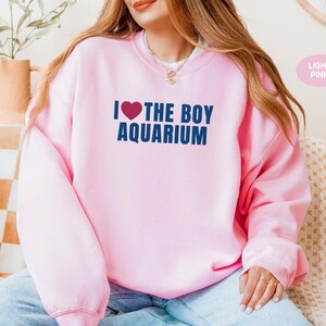 May include: Light pink sweatshirt with the text "I ❤️ THE BOY AQUARIUM" in navy blue. The heart symbol is red. The sweatshirt is a crew neck style and is made of a soft material. The text is centered on the front of the sweatshirt.