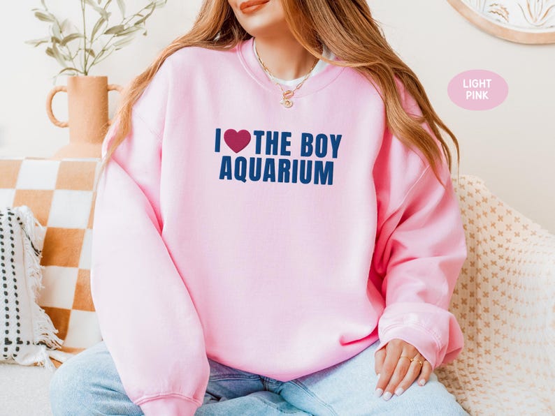 May include: Light pink sweatshirt with the text "I ❤️ THE BOY AQUARIUM" in navy blue. The heart symbol is red. The sweatshirt is a crew neck style and is made of a soft material. The text is centered on the front of the sweatshirt.