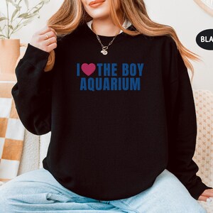 May include: Black sweatshirt with the text "I &hearts; THE BOY AQUARIUM" in blue and a pink heart. The sweatshirt is a crew neck style and is made of a soft material. The word "BLACK" is in a black oval.