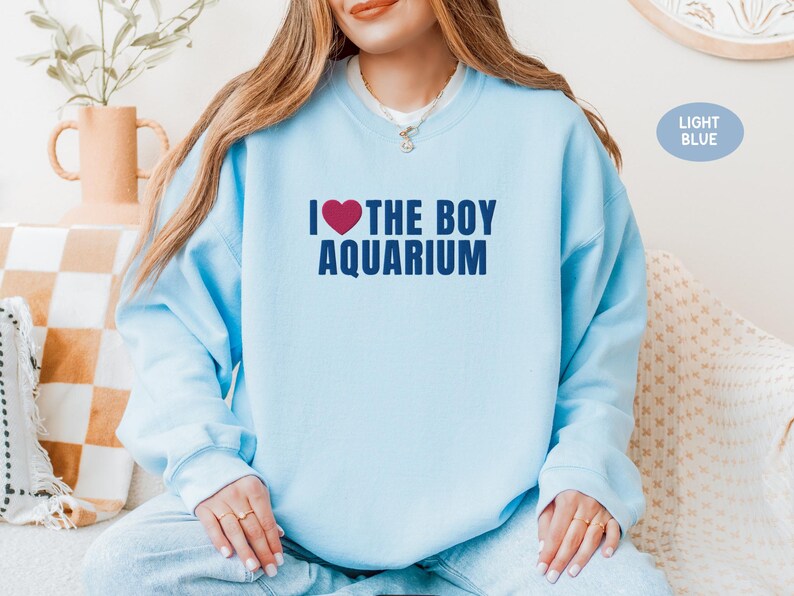 May include: Light blue sweatshirt with the text "I ❤️ THE BOY AQUARIUM" in navy blue and a red heart. The sweatshirt is a solid light blue color. The image also includes a light blue color swatch.