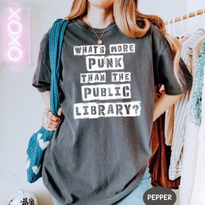 Op de afbeelding: Donkergrijs t-shirt met de witte tekst "WHAT'S MORE PUNK THAN THE PUBLIC LIBRARY?". Het shirt heeft een relaxte pasvorm, ideaal voor casual wear. Een blauwe tas is zichtbaar aan de linkerkant van de afbeelding.
