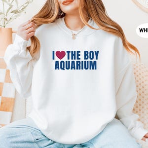 May include: White sweatshirt with the text "I ❤️ THE BOY AQUARIUM" in navy blue and a red heart. The sweatshirt is a casual style, suitable for everyday wear. The image shows a person wearing the sweatshirt.