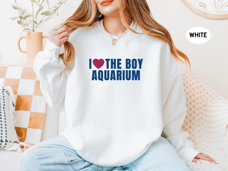 May include: White sweatshirt with the text "I ❤️ THE BOY AQUARIUM" in navy blue and a red heart. The sweatshirt is a casual style, suitable for everyday wear. The image shows a person wearing the sweatshirt.
